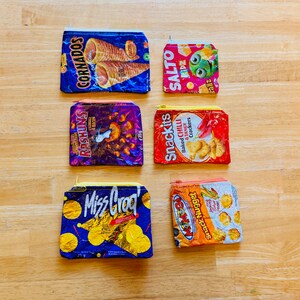 May include: Six colorful zippered pouches made from recycled snack food packaging. The pouches feature various snack brands including Cornaros, Rashuns, Snackits, Miss Grog, Snack Nugget, and Salto.