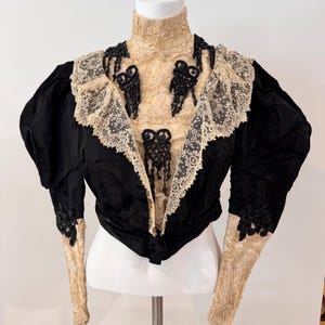 May include: A vintage black velvet jacket with a high collar and long sleeves. The jacket features intricate lace and beadwork details in cream and black. The sleeves are made of cream-colored lace.