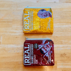 May include: Two small zippered pouches made from recycled food packaging. The pouches are both yellow and red with white text. The text on the yellow pouch reads "REAL HAND COOKED HAM & ENGLISH MUSTARD WITH BAGS OF CHARACTER". The text on the red pouch reads "REAL HAND COOKED ROAST OX great taste WITH BAGS OF CHARACTER".
