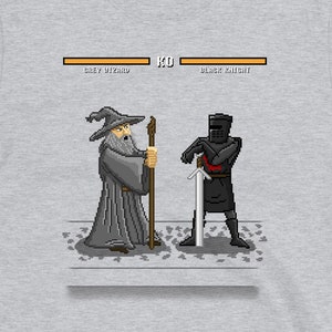 May include: A light gray t-shirt featuring a pixelated graphic of a Grey Wizard facing off against a Black Knight. The design includes a health bar labeled "Grey Wizard" and "Black Knight" with the word "KO" in the middle. The website address www.paulsimic.com is at the bottom.
