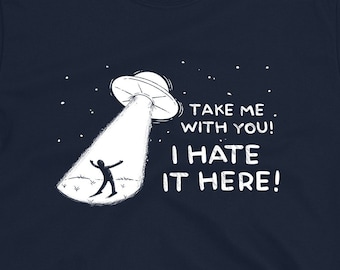 May include: Navy blue t-shirt with a humorous graphic of a person being beamed up by a UFO. The text reads "Take me with you! I hate it here!" in white. The design includes a starry sky and a grassy field.