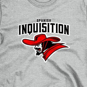 Puede incluir: Camiseta gris claro con las palabras "SPANISH INQUISITION" en blanco y negro, y un gráfico rojo y negro de un hombre con sombrero. La dirección del sitio web "WWW.PAULSIMIC.COM" está en la parte inferior.