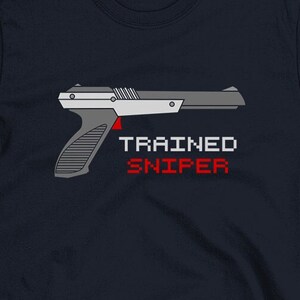 May include: A navy blue t-shirt featuring a pixelated image of a NES Zapper gun and the text "TRAINED SNIPER" in red and white.