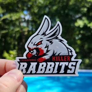 Può includere: Un adesivo raffigurante un coniglio cartone animato minaccioso con occhi rossi e sangue intorno alla bocca. Il testo "KILLER RABBITS" è visualizzato sotto il coniglio, con "CAERBANNOG" sopra. L'adesivo è tenuto contro uno sfondo sfocato.