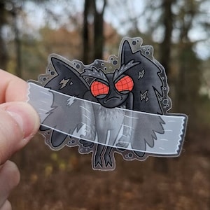 Puede incluir: Una pegatina transparente con una polilla de dibujos animados negra y gris con ojos rojos. La polilla está parcialmente cubierta con cinta adhesiva transparente. La pegatina tiene un fondo blanco con texto negro que dice "www.paulsimic.com".