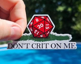May include: A white sticker with a red 20-sided die on a green patch of grass. The sticker says "Don't Crit On Me" and "www.paulsimic.com".