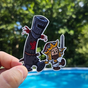 Könnte beinhalten: Ein Aufkleber mit Cartoon-Illustrationen von zwei mittelalterlichen Charakteren. Einer ist ein schwarzer Ritter mit Blutspritzern, der andere ein König mit einem Schwert. Der Aufkleber wird vor einem Pool gehalten. Die Website www.paulsimic.com ist sichtbar.