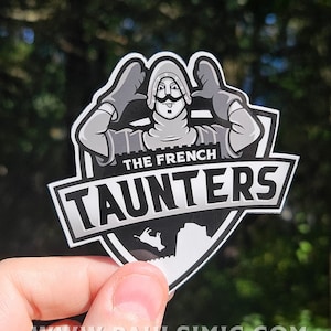 Op de afbeelding: Een sticker met een cartoon illustratie van een Franse soldaat met een grote snor en handschoenen. De tekst "THE FRENCH TAUNTERS" wordt vetgedrukt weergegeven. Het ontwerp van de sticker is voornamelijk zwart, wit en grijs, met een schildachtige vorm.