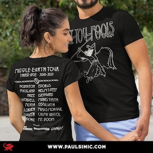 May include: A black t-shirt featuring a white graphic of a wizard with a staff and the text FLY YOU FOOLS on the front. The back displays a list of locations titled MIDDLE-EARTH TOUR THIRD AGE 3018-3019 with a chain graphic and text.