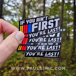 May include: A stack of rectangular stickers with a black background, featuring the white text IF YOU AIN'T FIRST YOU'RE LAST! and a vertical stripe design in yellow, red, and blue on the left side. The stickers have a glossy finish and bold lettering.