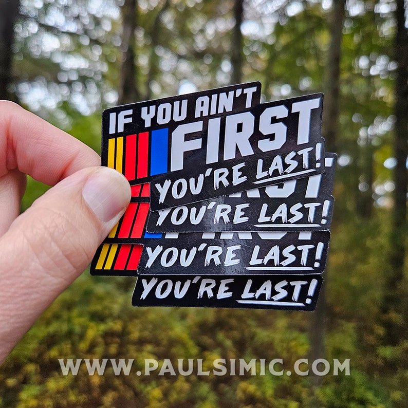 May include: A stack of rectangular stickers with a black background, featuring the white text IF YOU AIN'T FIRST YOU'RE LAST! and a vertical stripe design in yellow, red, and blue on the left side. The stickers have a glossy finish and bold lettering.