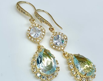 May include: A pair of gold-toned drop earrings. Each earring features a square, clear stone surrounded by smaller clear stones, connected to a larger, pear-shaped, light blue and yellow stone, also surrounded by smaller clear stones.