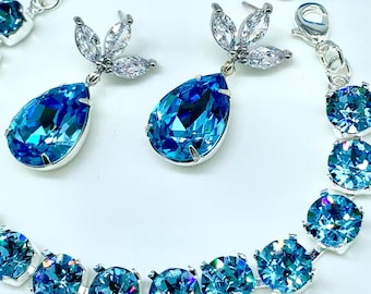 May include: A silver-toned bracelet and matching earrings set. The bracelet and earrings feature large, round and teardrop-shaped, faceted blue crystals. The earrings have a cluster of small, clear crystals at the top. The jewelry is set against a white background.