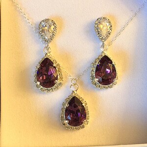 May include: A set of purple teardrop earrings and a matching necklace with silver settings and clear crystal accents. The jewelry is displayed in a white box with a purple ribbon.