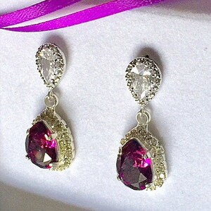 May include: A pair of dangle earrings with a pear-shaped purple gemstone surrounded by a halo of smaller clear stones. The earrings are silver-toned and have a smaller clear pear-shaped stone above the purple gemstone.