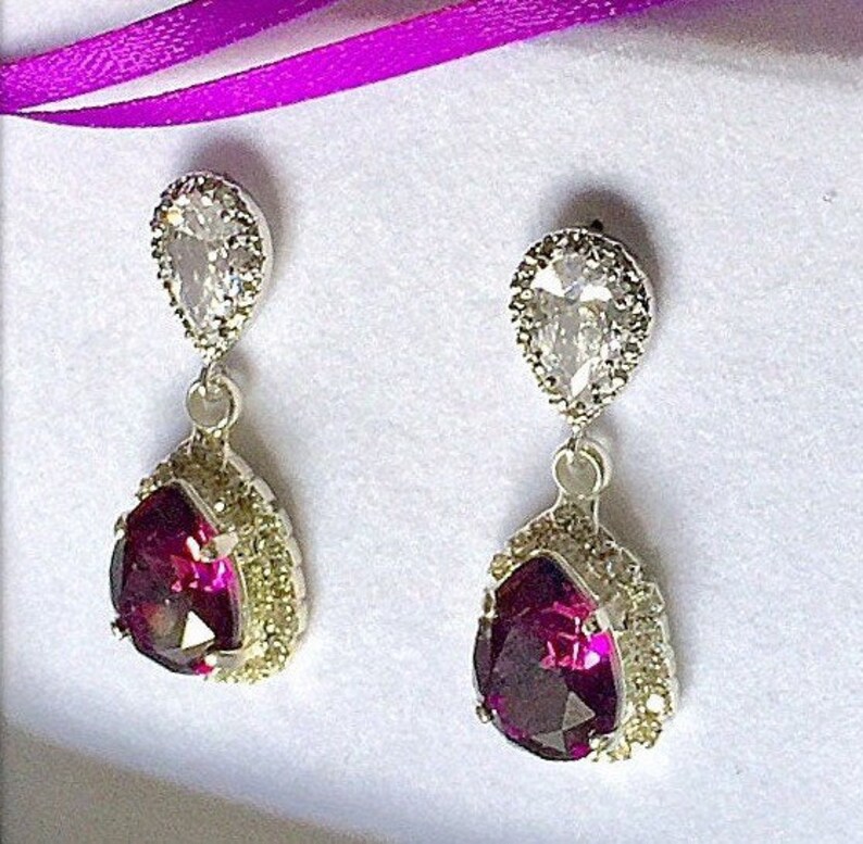 May include: A pair of dangle earrings with a pear-shaped purple gemstone surrounded by a halo of smaller clear stones. The earrings are silver-toned and have a smaller clear pear-shaped stone above the purple gemstone.