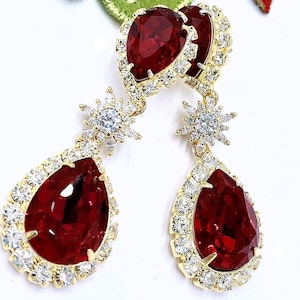 May include: A pair of gold-toned drop earrings featuring large, teardrop-shaped, deep red gemstones. The gemstones are surrounded by a halo of smaller, clear crystals. Additional starburst-shaped accents with clear crystals add to the earrings' sparkle.