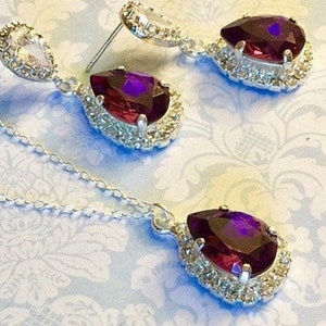 May include: A silver necklace and earrings set with a teardrop-shaped purple gemstone surrounded by clear rhinestones.