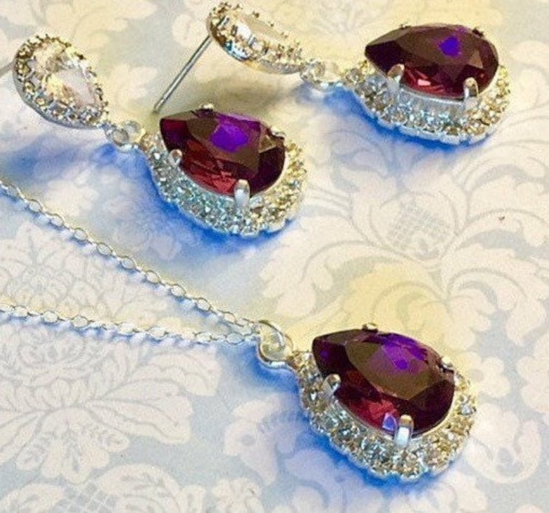 May include: A silver necklace and earrings set with a teardrop-shaped purple gemstone surrounded by clear rhinestones.