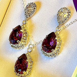 May include: A set of earrings and a necklace with a teardrop-shaped purple gemstone surrounded by a halo of smaller clear stones. The earrings and necklace are made of silver and have a delicate chain.