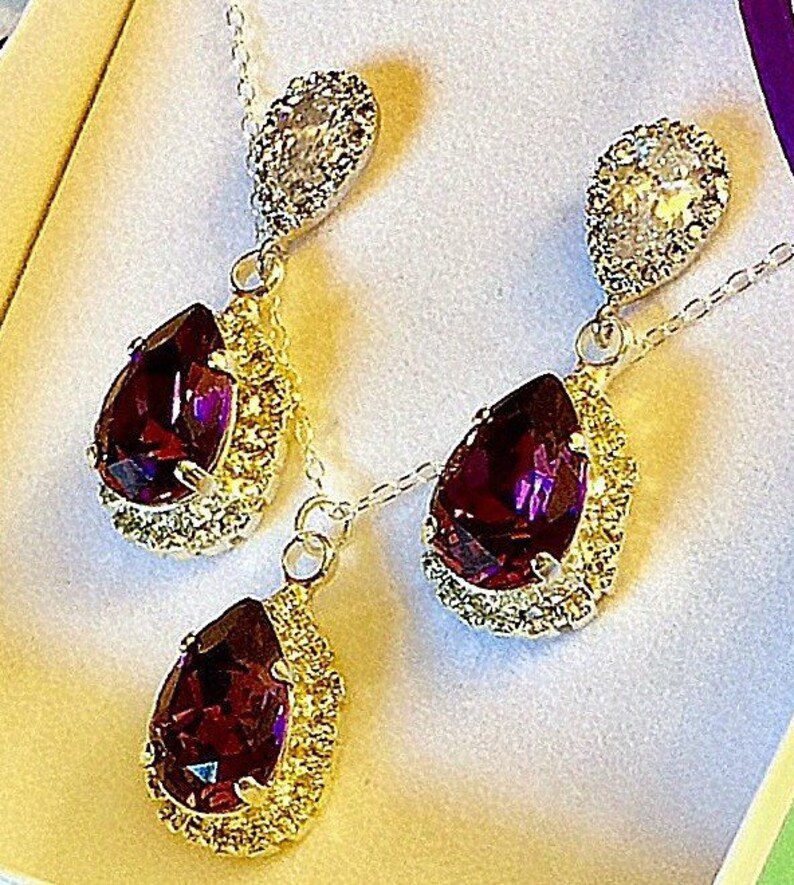 May include: A set of earrings and a necklace with a teardrop-shaped purple gemstone surrounded by a halo of smaller clear stones. The earrings and necklace are made of silver and have a delicate chain.