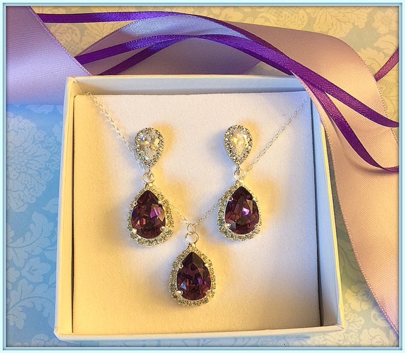 May include: A set of purple teardrop earrings and a matching necklace with silver settings and clear crystal accents. The jewelry is displayed in a white box with a purple ribbon.