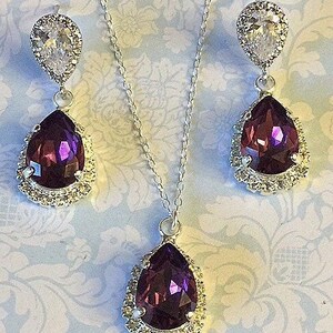 May include: A silver necklace and earrings set with a teardrop-shaped purple crystal surrounded by clear rhinestones.