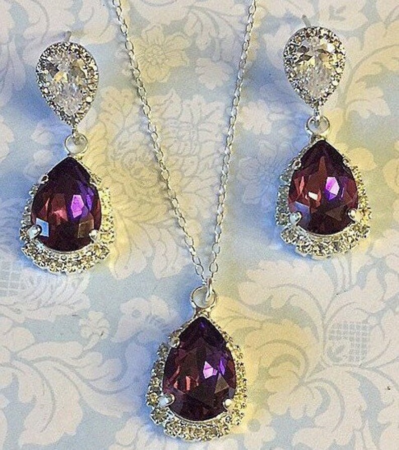 May include: A silver necklace and earrings set with a teardrop-shaped purple crystal surrounded by clear rhinestones.