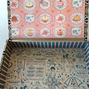 May include: A vintage-style wooden box with a hinged lid. The lid is decorated with a pink and white floral pattern and images of pastries. The inside of the box is lined with a blue and white striped fabric and features a black and white illustration of a Parisian bakery with a menu and a variety of pastries.