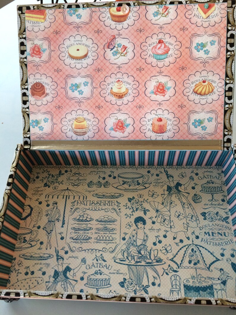May include: A vintage-style wooden box with a hinged lid. The lid is decorated with a pink and white floral pattern and images of pastries. The inside of the box is lined with a blue and white striped fabric and features a black and white illustration of a Parisian bakery with a menu and a variety of pastries.