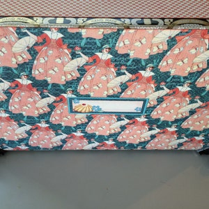 May include: A teal blue box with a pink and white floral pattern featuring women in dresses and parasols. The box has a label that says "Hand Made".