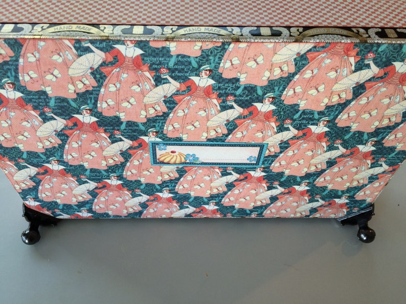 May include: A teal blue box with a pink and white floral pattern featuring women in dresses and parasols. The box has a label that says "Hand Made".