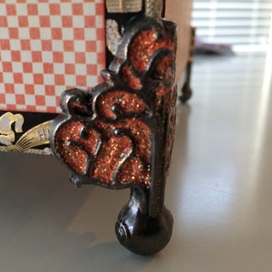 May include: A decorative black metal foot with an intricate design and a red glitter finish. The foot is round and has a ball-shaped base.
