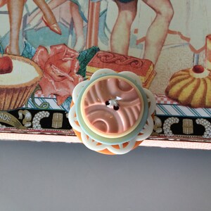 May include: A close-up of a pastel blue and white button with a pink and orange center. The button has a decorative design and is surrounded by a white border. The button is on a background of a craft project with a hand-made label.