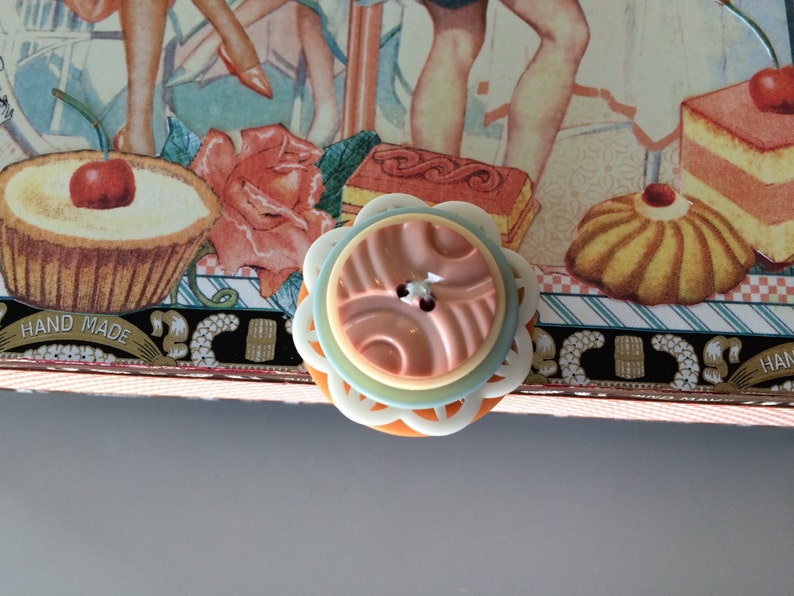 May include: A close-up of a pastel blue and white button with a pink and orange center. The button has a decorative design and is surrounded by a white border. The button is on a background of a craft project with a hand-made label.