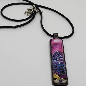 May include: A black cord necklace with a rectangular pendant featuring a cityscape with pink and purple hues. The pendant has a silver frame.