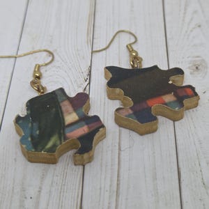 May include: A pair of wooden puzzle piece earrings. The earrings have a gold-colored hook and are painted with a colorful abstract design. The wood has a natural, light brown color.