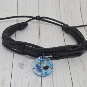 May include: Black leather cord bracelet with a round pendant. The pendant is clear with blue and black accents, including glitter and shapes. The bracelet is adjustable.