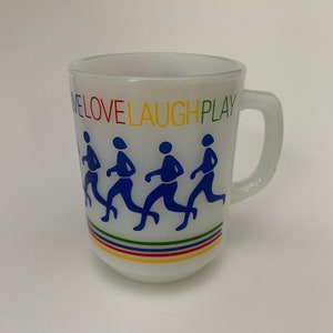 Może przedstawiać: Biały kubek ceramiczny z wzorem w niebieskie, czerwone, żółte i zielone paski. Kubek ma uchwyt i napis "We Love Laugh Play" z niebieskimi postaciami biegnącymi.