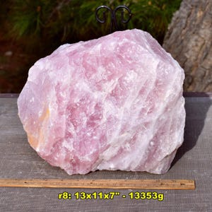May include: A large, rough-hewn rose quartz crystal. The stone is a soft pink color with white veining. The dimensions are 13x11x7 inches. The text "r8: 13x11x7" - 13353g" is visible.