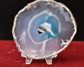 May include: A translucent blue and white agate slice with a central geode opening, resting on a clear acrylic stand. The stone features concentric banding and measures 12.7x12.7 cm with a weight of 131 g as indicated by the text 62: 5x5 - 131 g.