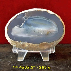 May include: A polished agate stone, displaying bands of gray, white, and blue hues. The stone is oval-shaped and rests on a clear plastic stand. The text on the image reads: 19: 4x3x.5" - 263 g.