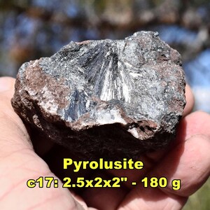 May include: A rough, dark gray pyrolusite mineral specimen with a radial, crystalline structure. The rock measures 2.5 x 2 x 2 inches and weighs 180 grams. The text "Pyrolusite" and other details are visible in yellow.