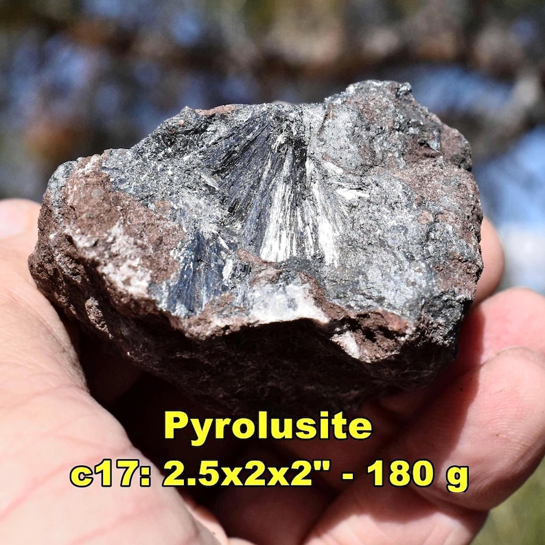 May include: A rough, dark gray pyrolusite mineral specimen with a radial, crystalline structure. The rock measures 2.5 x 2 x 2 inches and weighs 180 grams. The text "Pyrolusite" and other details are visible in yellow.