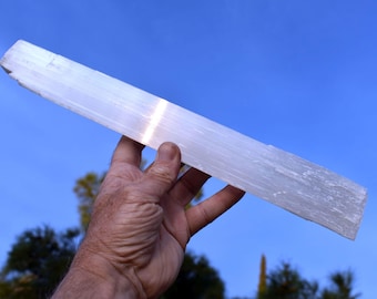 May include: A large, white selenite crystal wand with a smooth, polished surface. The crystal is held in a hand against a blue sky background.