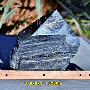 May include: A large, dark, crystalline mineral specimen with a pointed top and layered, fibrous texture. A wooden ruler placed in front shows the object measures 9 inches by 8 inches by 3 inches, with a weight of 3280 grams as noted in text.