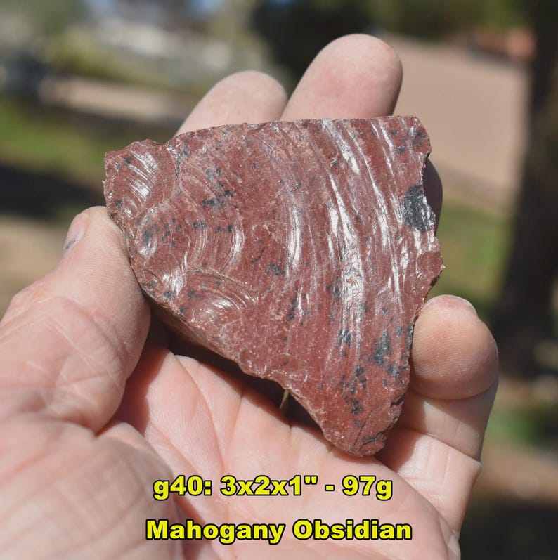 May include: A piece of mahogany obsidian, a reddish-brown volcanic glass, is held in a hand. The stone measures approximately 3 x 2 x 1 inches and weighs 97 grams. The surface displays swirling patterns and dark inclusions.