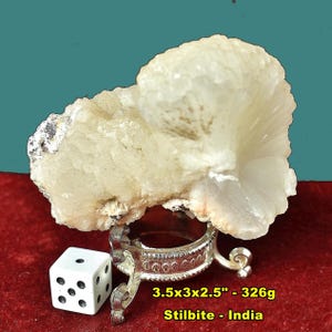 May include: A cream-colored stilbite crystal cluster, 3.5 x 3 x 2.5 inches, sits on a decorative silver stand. A white die with black dots is next to the stand. The text reads "Stilbite - India".
