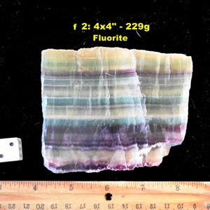 May include: A rectangular slab of banded fluorite mineral featuring horizontal layers of purple, green, yellow, and white. The text f 2: 4x4" - 229g Fluorite is printed above the stone. A white die and a wooden ruler are visible for scale.