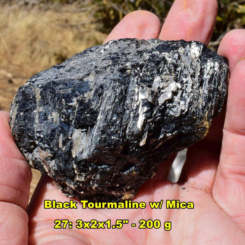 May include: A rough, black tourmaline stone with visible mica inclusions. The stone is held in a hand, showcasing its dark, textured surface. The text on the image reads "Black Tourmaline w/ Mica" and "27: 3x2x1.5" - 200 g".